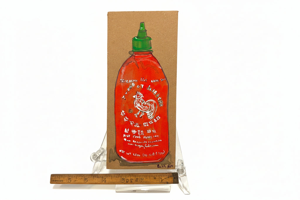 The Rachel Nelson • Rooster Sauce by Thyne Goods is a cardboard cutout painted with a red Sriracha bottle, displayed upright on a clear plastic stand and shown next to a wooden ruler for scale.