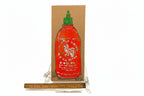 The Rachel Nelson • Rooster Sauce by Thyne Goods is a cardboard cutout painted with a red Sriracha bottle, displayed upright on a clear plastic stand and shown next to a wooden ruler for scale.