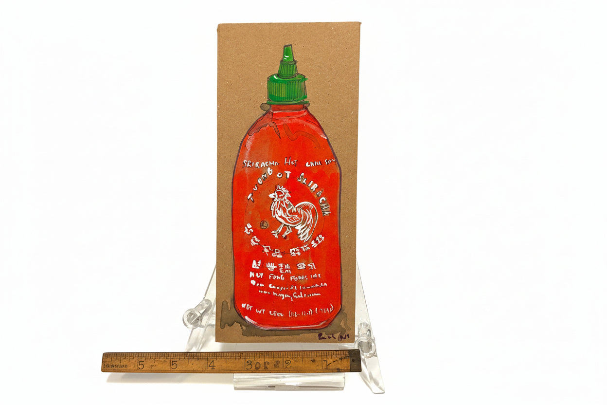 The Rachel Nelson • Rooster Sauce by Thyne Goods is a cardboard cutout painted with a red Sriracha bottle, displayed upright on a clear plastic stand and shown next to a wooden ruler for scale.