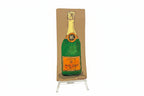 A rectangular cardboard piece titled "Rachel Nelson • Champagne" by Thyne Goods, featuring a painted Veuve Clicquot bottle, stands upright on a clear display against a white background, evoking Rachel Nelson’s immersive art style.