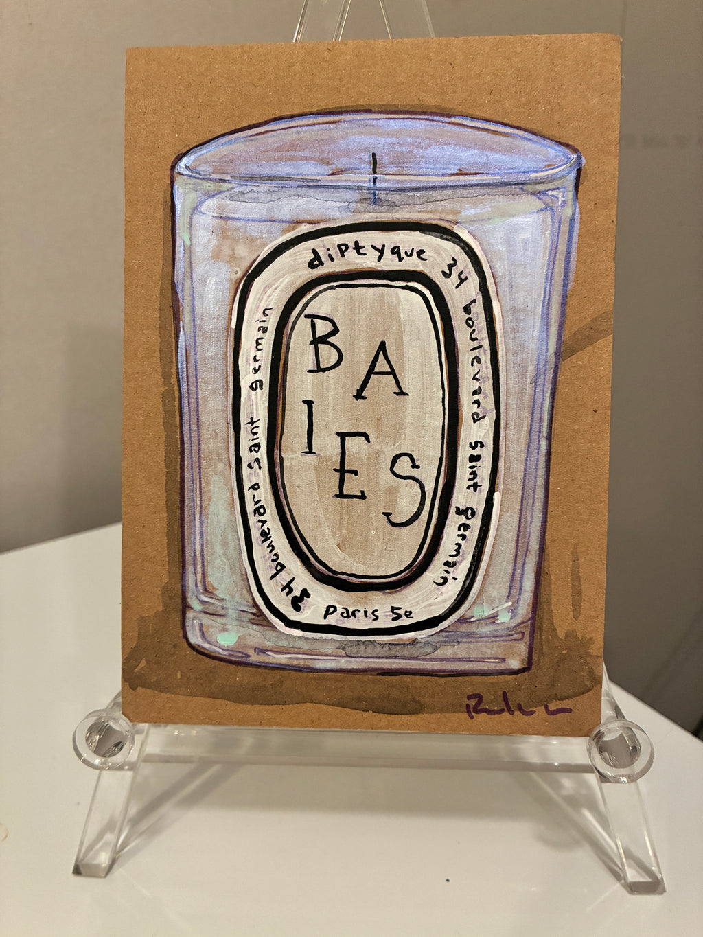 A hand-painted Rachel Nelson • Baies Paris candle illustration by Thyne Goods on brown paper, set on a clear stand. The detailed label contrasts with the signed purple artwork in the bottom right corner.