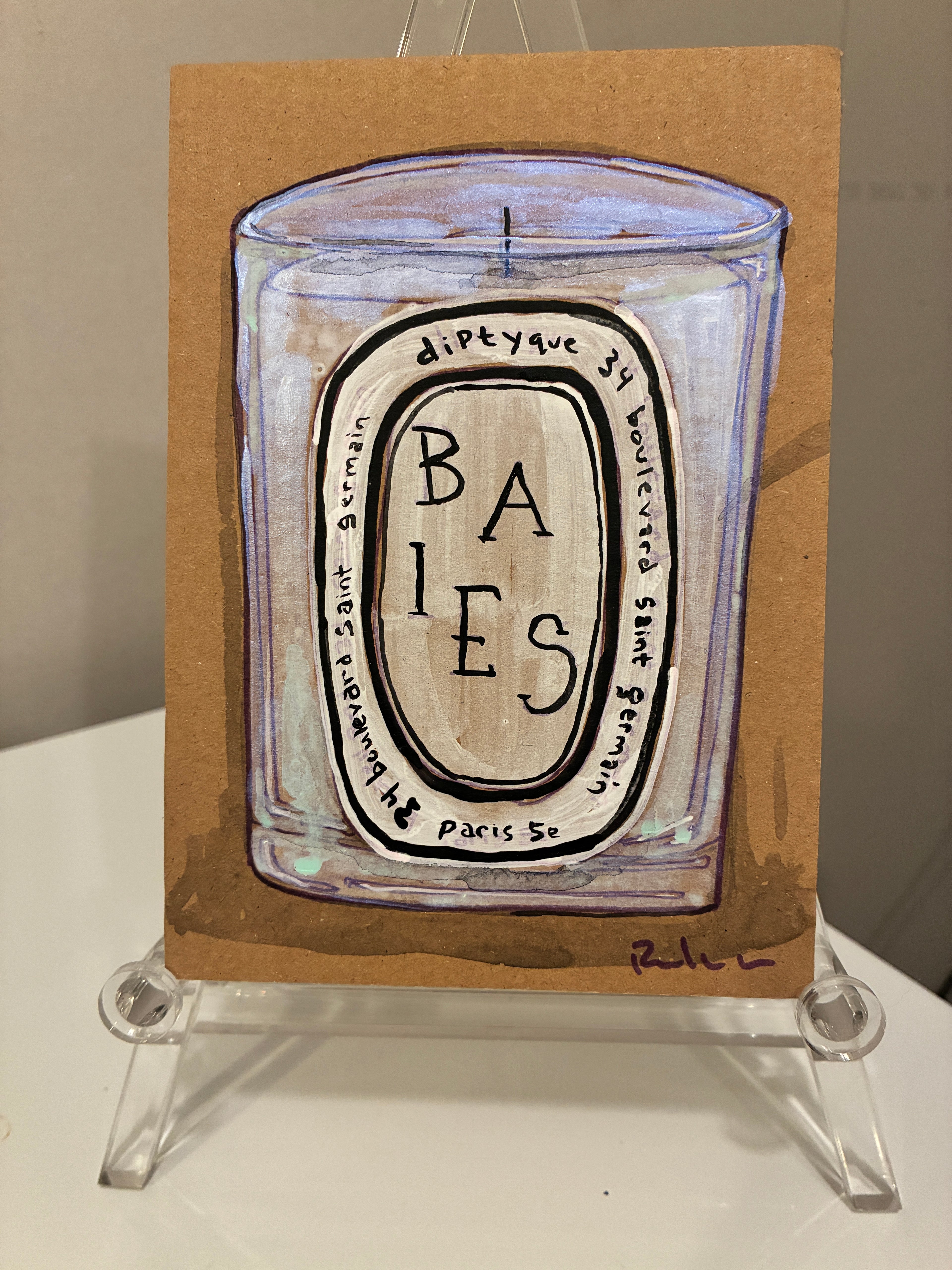 A hand-painted Rachel Nelson • Baies Paris candle illustration by Thyne Goods on brown paper, set on a clear stand. The detailed label contrasts with the signed purple artwork in the bottom right corner.