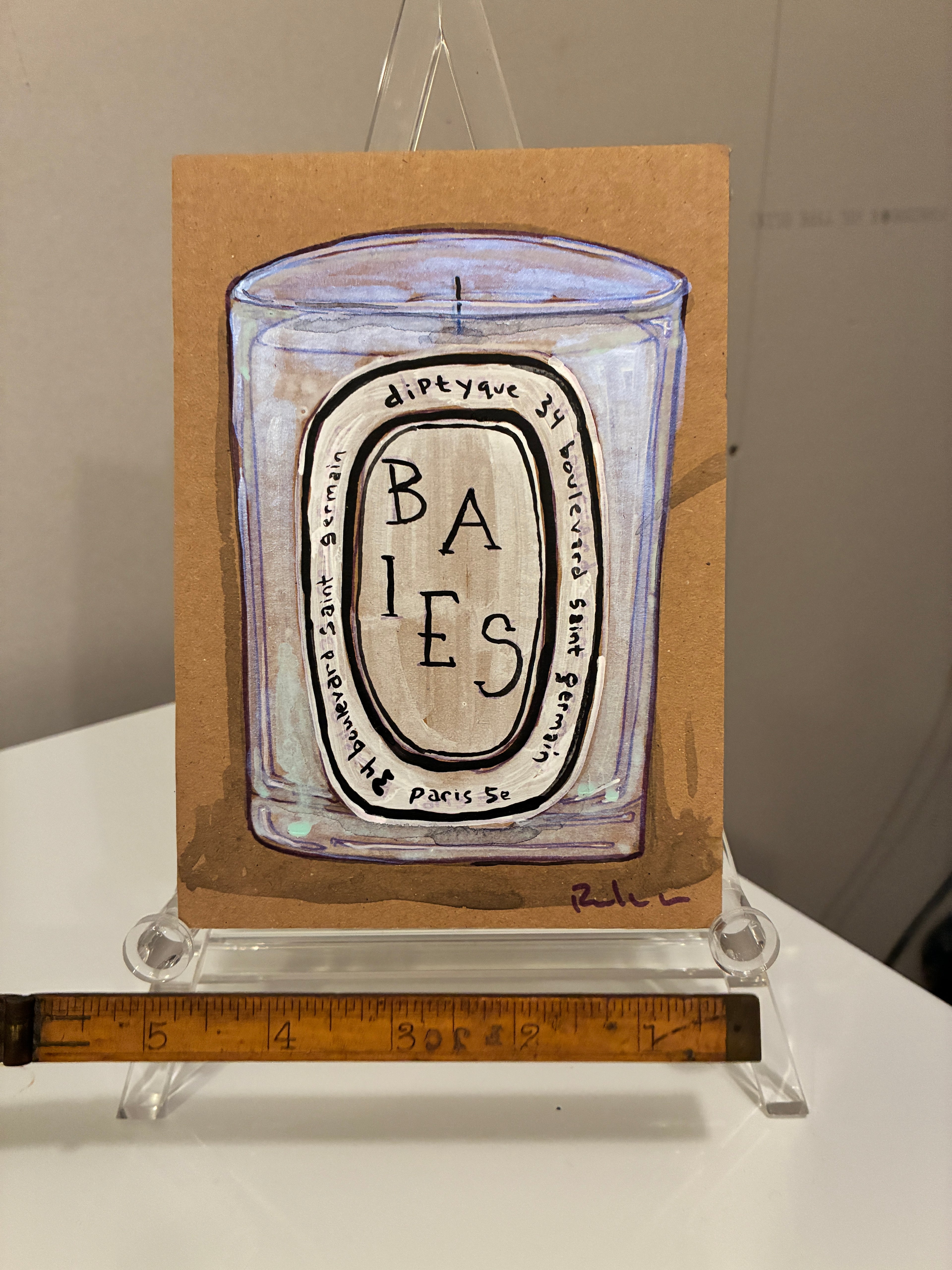A drawing of the Rachel Nelson • Baies Paris candle by Thyne Goods is shown on brown card with a clear stand. A yellow ruler sits in front, set against a white surface and beige wall in the background.