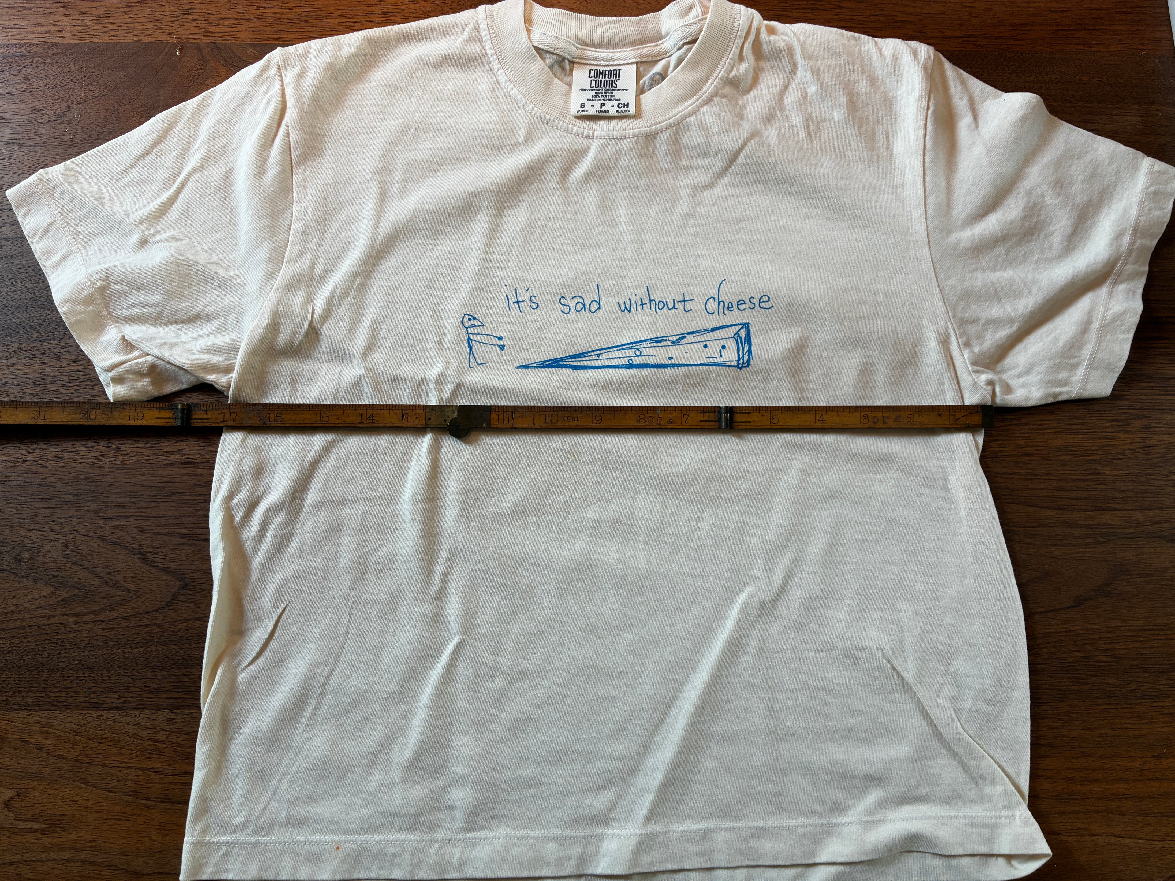 Thyne Goods' limited-edition Cheese cropped tee features a blue cheese wedge graphic and "Its sad without cheese." Printed on 100% cotton, it comes with a wooden ruler displayed horizontally across the front.
