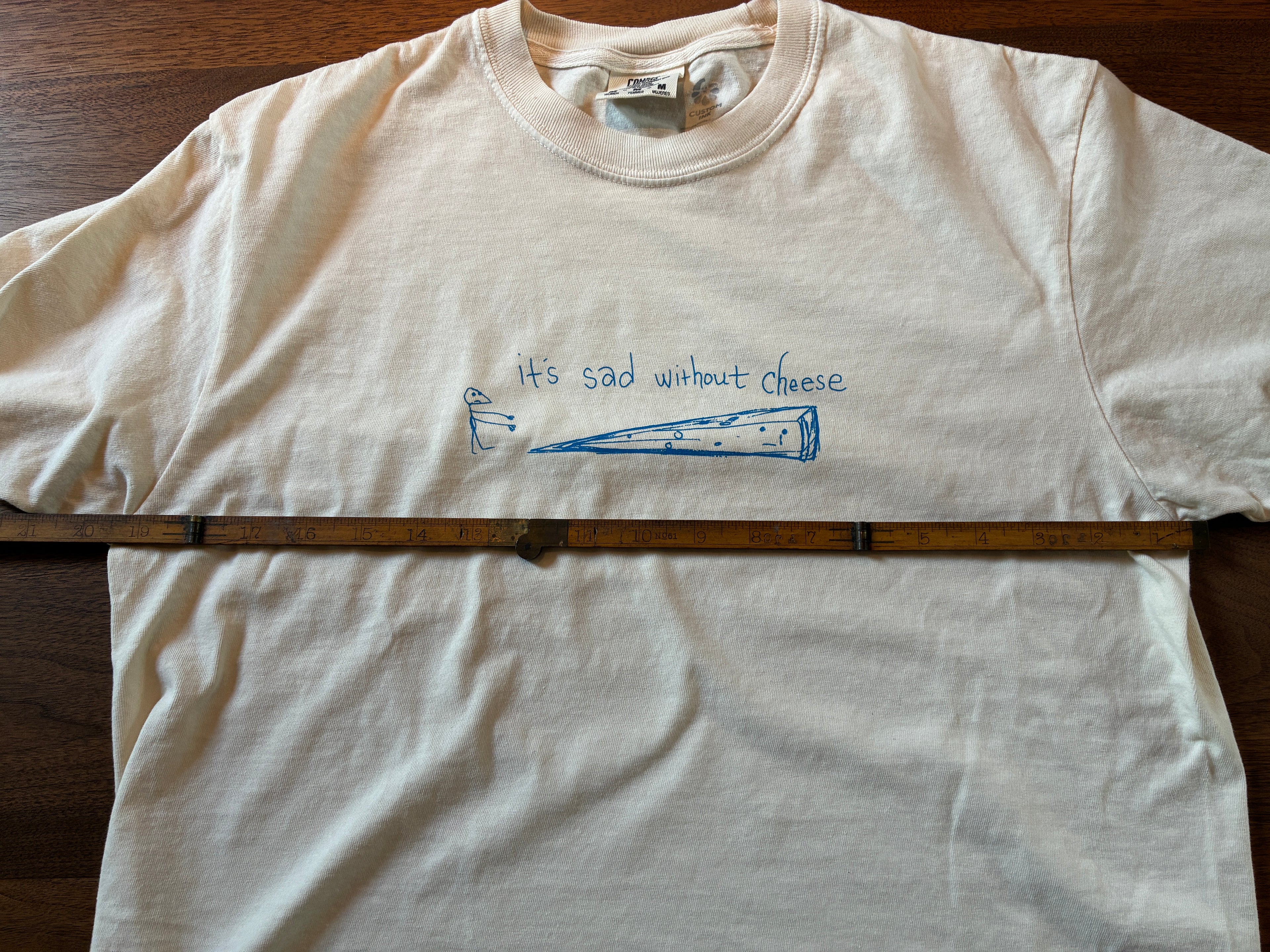 Thyne Goods’ limited edition “Cheese” cropped tee is 100% cotton, features a blue cheese wedge with a sad face and "It's sad without cheese" text on white fabric. A ruler is placed horizontally across the shirt in the image.