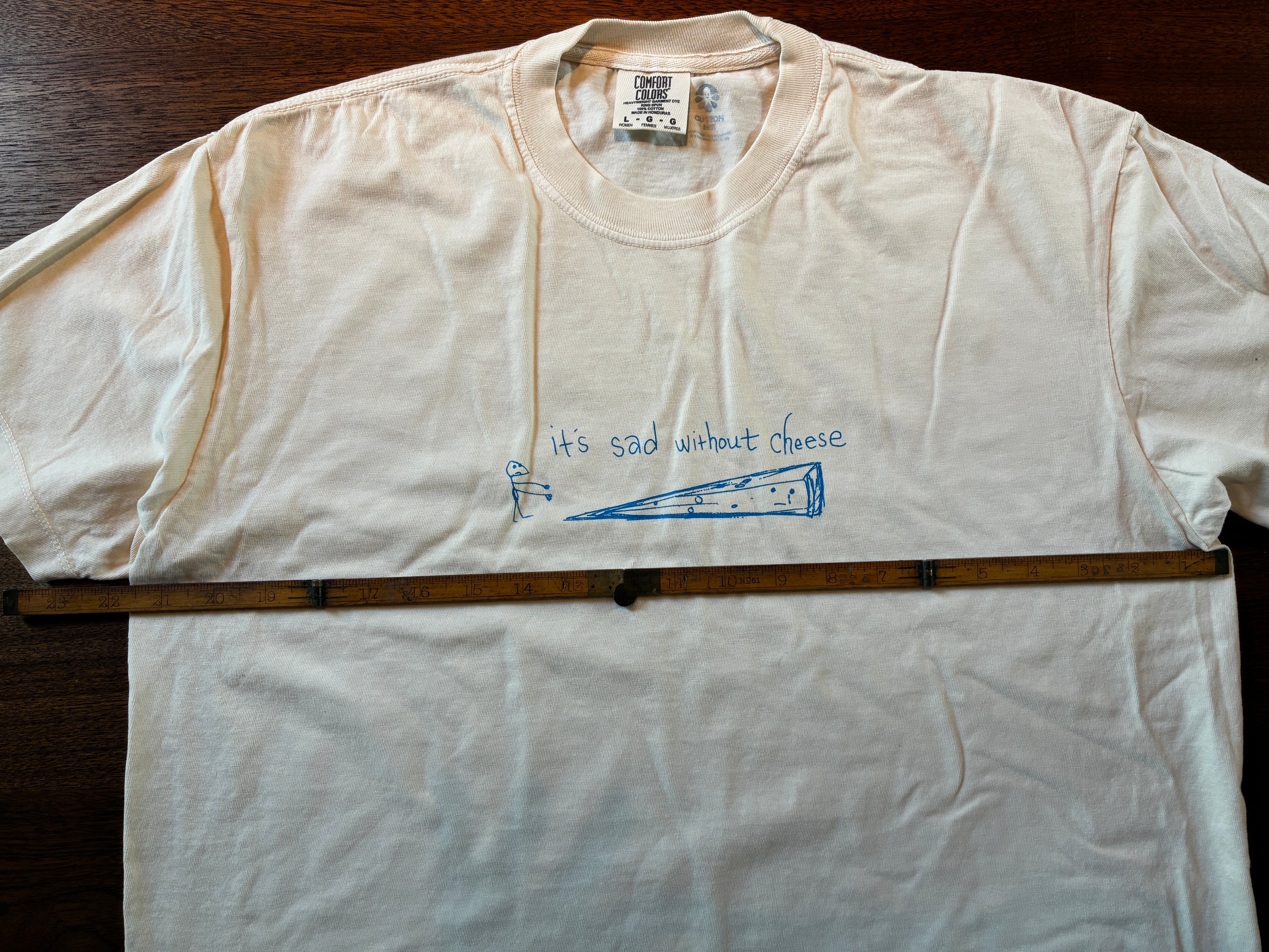 The Thyne Goods 'Cheese' tee is a cream, 100% cotton shirt featuring a simple blue sad mouse drawing facing cheese, with "It's sad without cheese" on the chest. A wooden ruler rests atop this limited edition design.