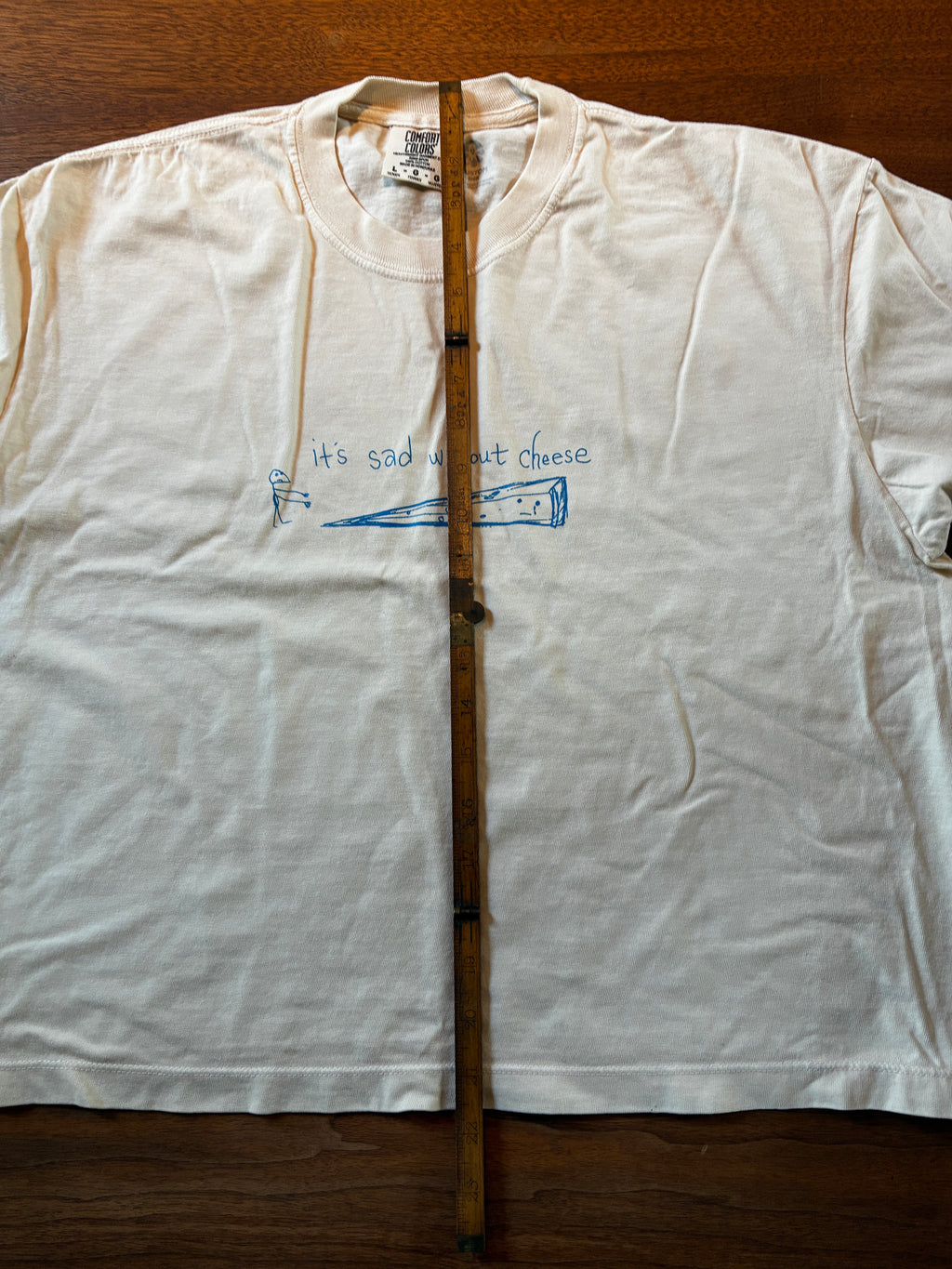 The Cheese cropped tee by Thyne Goods features a cream cotton design with simple blue wedge and mouse art, the phrase "it’s sad without cheese," and a wooden ruler detail. This is a limited edition release.