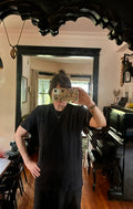A person wearing the Thyne Goods Baja Tee Man Sized takes a mirror selfie in a room with wooden floors, a piano, black-framed mirror, and various decorations and furniture in the background.