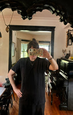 A person wearing the Thyne Goods Baja Tee Man Sized takes a mirror selfie in a room with wooden floors, a piano, black-framed mirror, and various decorations and furniture in the background.