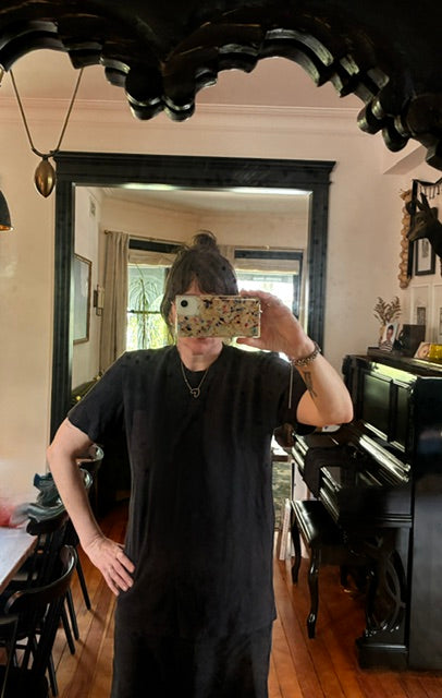 A person wearing the Thyne Goods Baja Tee Man Sized takes a mirror selfie in a room with wooden floors, a piano, black-framed mirror, and various decorations and furniture in the background.