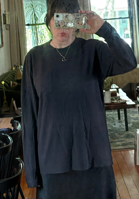 Person taking a mirror selfie in a dimly lit room, wearing a loose-fitting Thyne Goods Baja Long Sleeve Tee and a heart pendant necklace. Background features a living area with wooden floors and furniture.