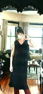 Wearing a Thyne Goods Basic Cami Tank in black, a person takes a mirror selfie in a living room with large windows, beige curtains, a green couch, and a wooden coffee table visible in the background.