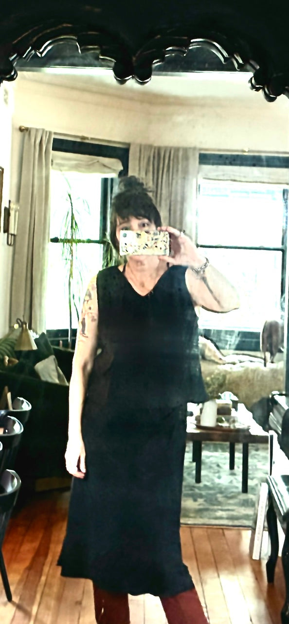 Wearing a Thyne Goods Basic Cami Tank in black, a person takes a mirror selfie in a living room with large windows, beige curtains, a green couch, and a wooden coffee table visible in the background.