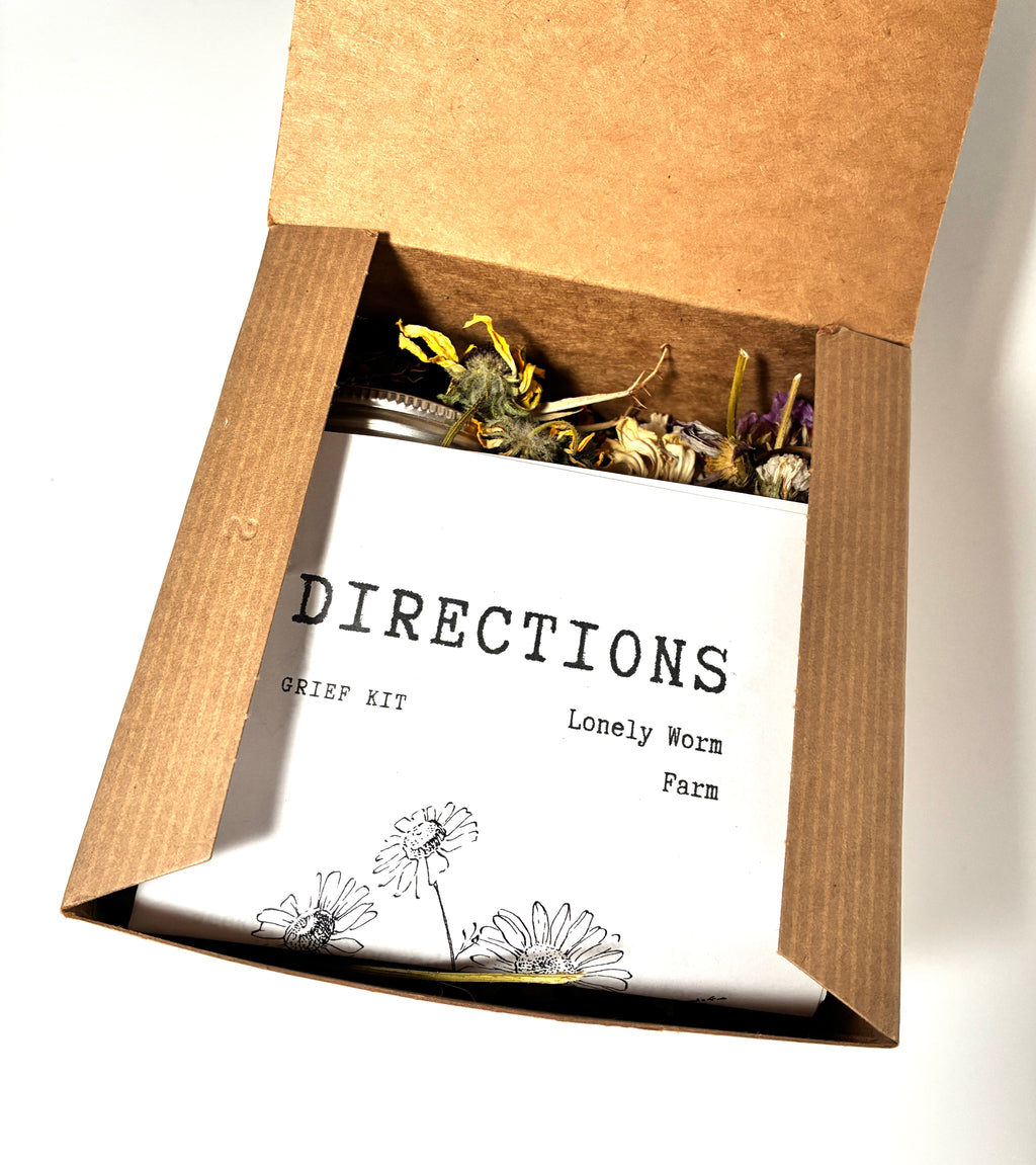 An open cardboard box displays the Grief Kit from Lonely Worm Farm, featuring a booklet with hand-drawn flowers and dried herbs, making it a thoughtful comfort or remembrance gift for someone experiencing loss.