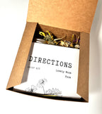 An open cardboard box displays the Grief Kit from Lonely Worm Farm, featuring a booklet with hand-drawn flowers and dried herbs, making it a thoughtful comfort or remembrance gift for someone experiencing loss.