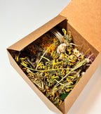 An open cardboard box containing a glass jar nestled among dried yellow and white flowers, viewed from above on a white background—this Grief Kit from Lonely Worm Farm offers a thoughtful gift for those experiencing loss.