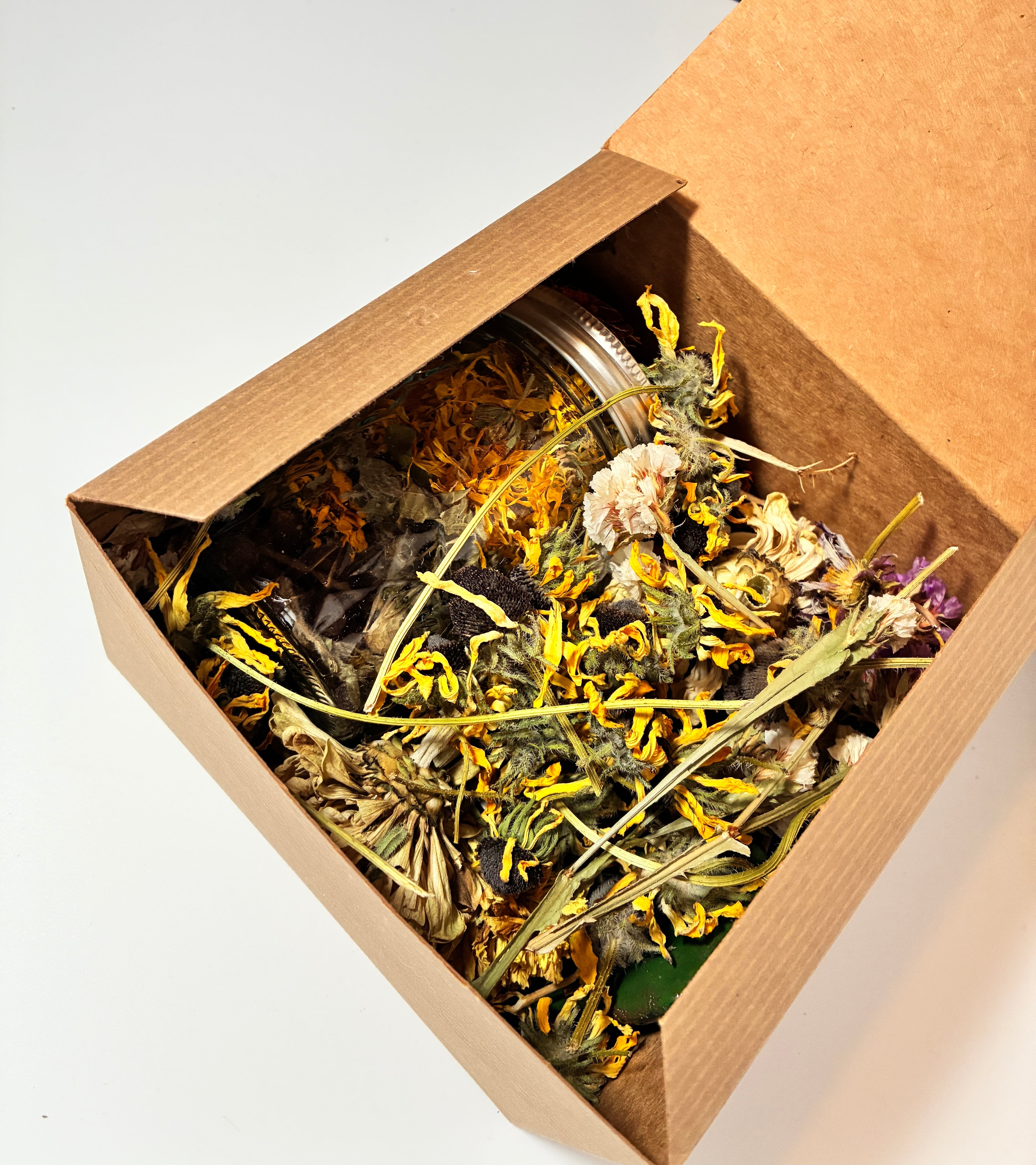 An open cardboard box containing a glass jar nestled among dried yellow and white flowers, viewed from above on a white background—this Grief Kit from Lonely Worm Farm offers a thoughtful gift for those experiencing loss.