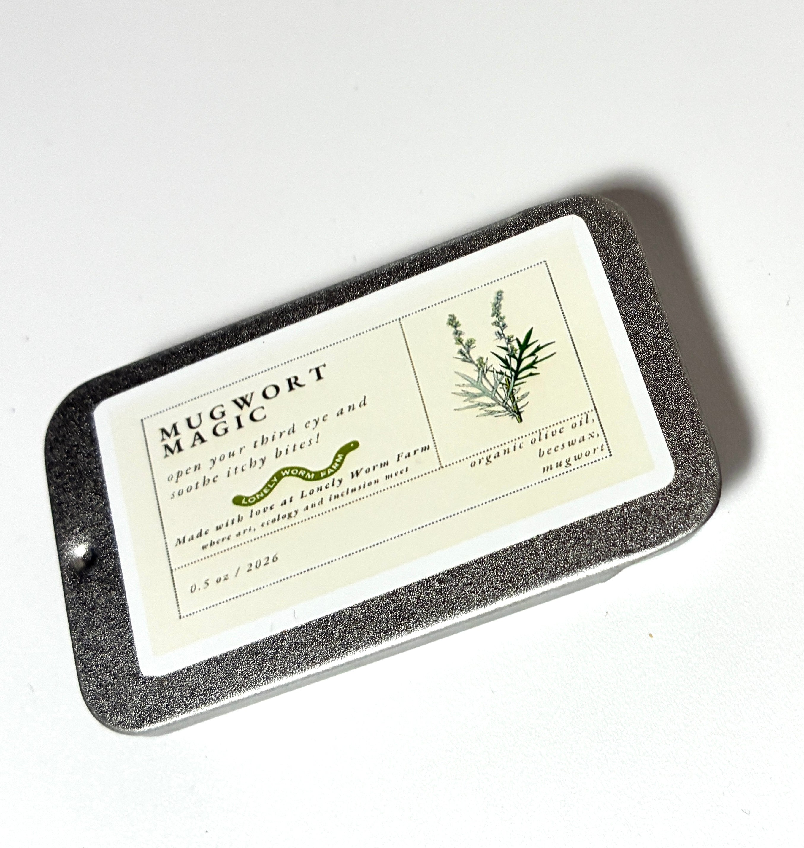 A rectangular tin labeled “Mugwort Magic from Lonely Worm Farm” by Thyne Goods features an illustration of mugwort. The light green and white label highlights the product’s eye and sleep benefits, with organic ingredients like mugwort and organic olive oil.