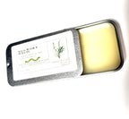A rectangular metal tin from Thyne Goods labeled “Mugwort Magic from Lonely Worm Farm” features a white label with a small plant illustration. The lid is partially opened, revealing the pale yellow balm inside, crafted with organic olive oil—perfect for soothing itchy bite relief.