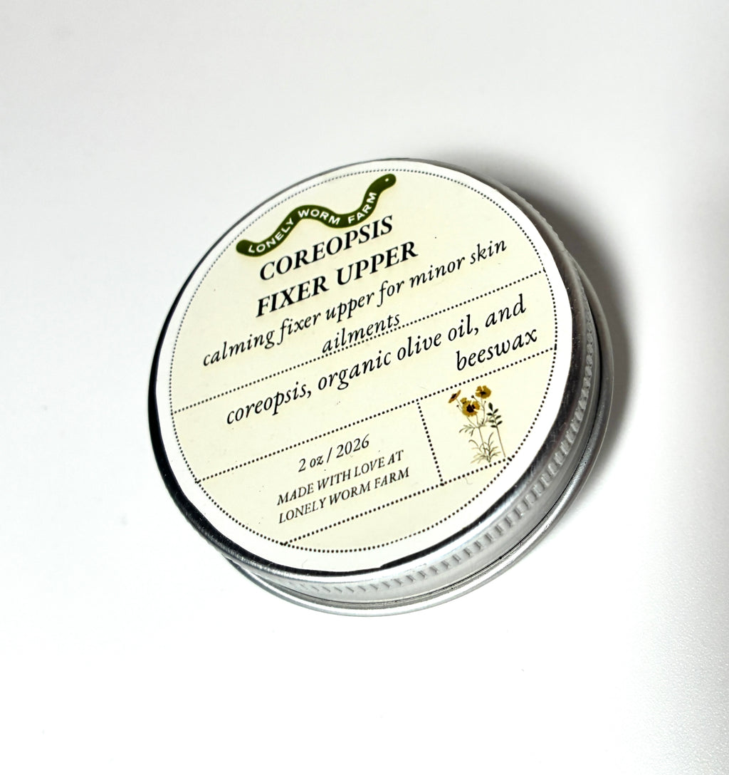 A round tin labeled Coreopsis Fixer Upper from Lonely Worm Farm by Thyne Goods, formulated with coreopsis, organic olive oil, and beeswax, serves as a calming skin fixer for minor skin ailments.