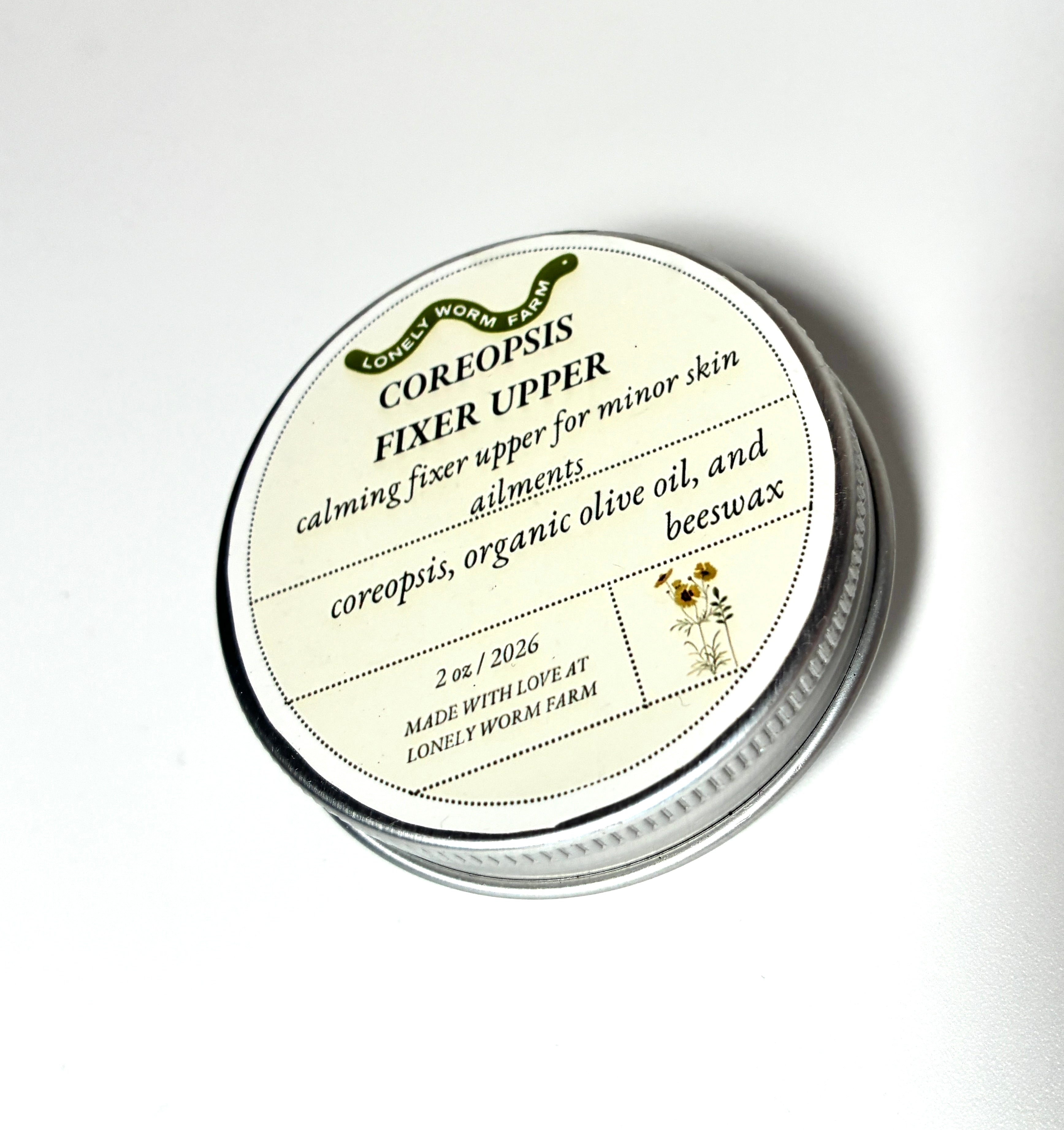 A round tin labeled Coreopsis Fixer Upper from Lonely Worm Farm by Thyne Goods, formulated with coreopsis, organic olive oil, and beeswax, serves as a calming skin fixer for minor skin ailments.