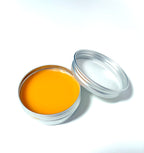 A small, round metal tin of Coreopsis Fixer Upper from Lonely Worm Farm by Thyne Goods—filled with smooth, orange balm made with organic olive oil to soothe minor skin ailments—rests on a white surface with its lid beside it.