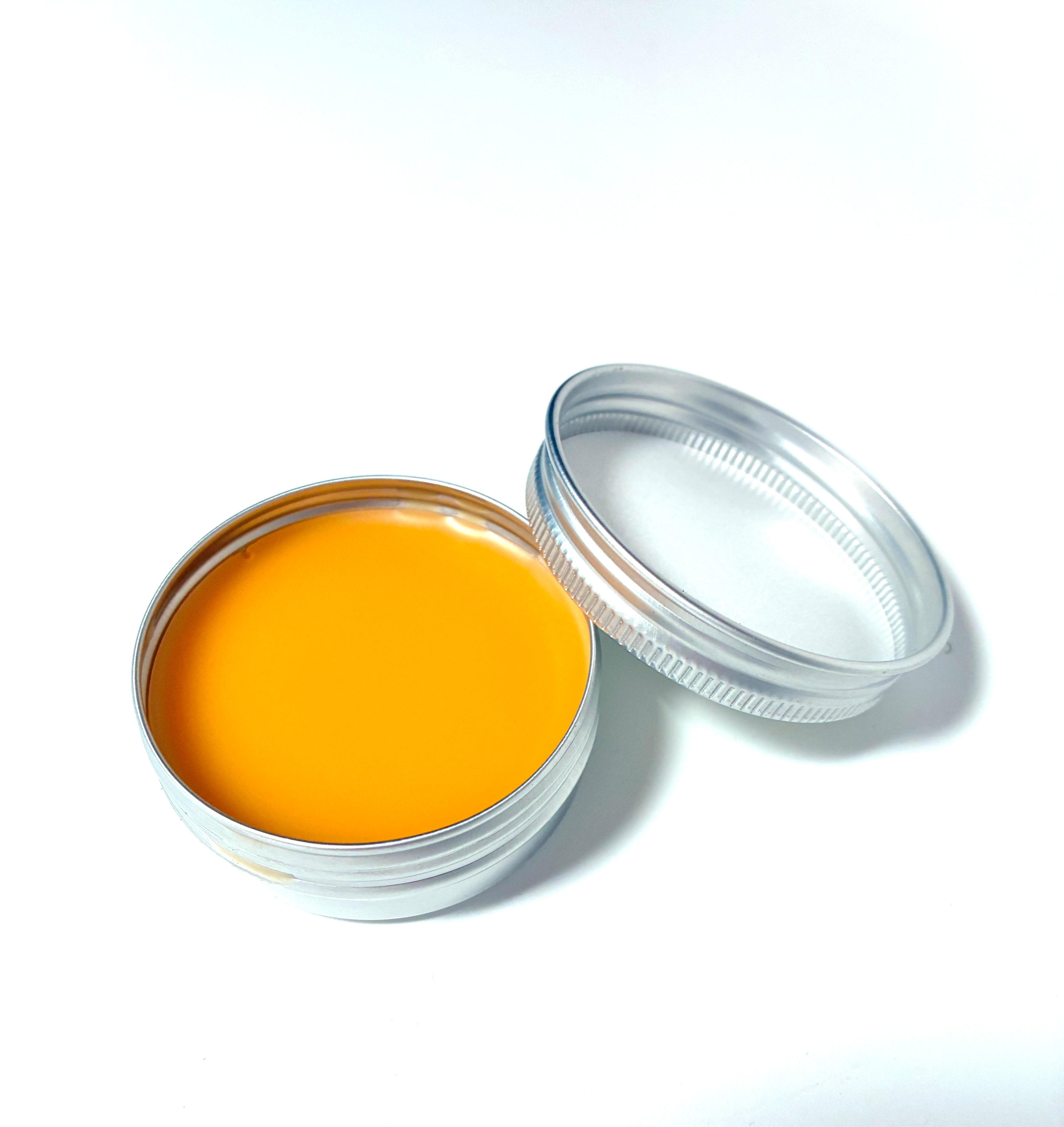 A small, round metal tin of Coreopsis Fixer Upper from Lonely Worm Farm by Thyne Goods—filled with smooth, orange balm made with organic olive oil to soothe minor skin ailments—rests on a white surface with its lid beside it.
