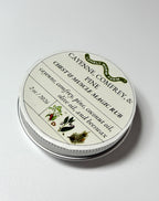 A round tin of Cayenne, Comfrey, and Pine Balm from Lonely Worm Farm by Thyne Goods, adorned with ingredient details and botanical illustrations, rests on a white surface.