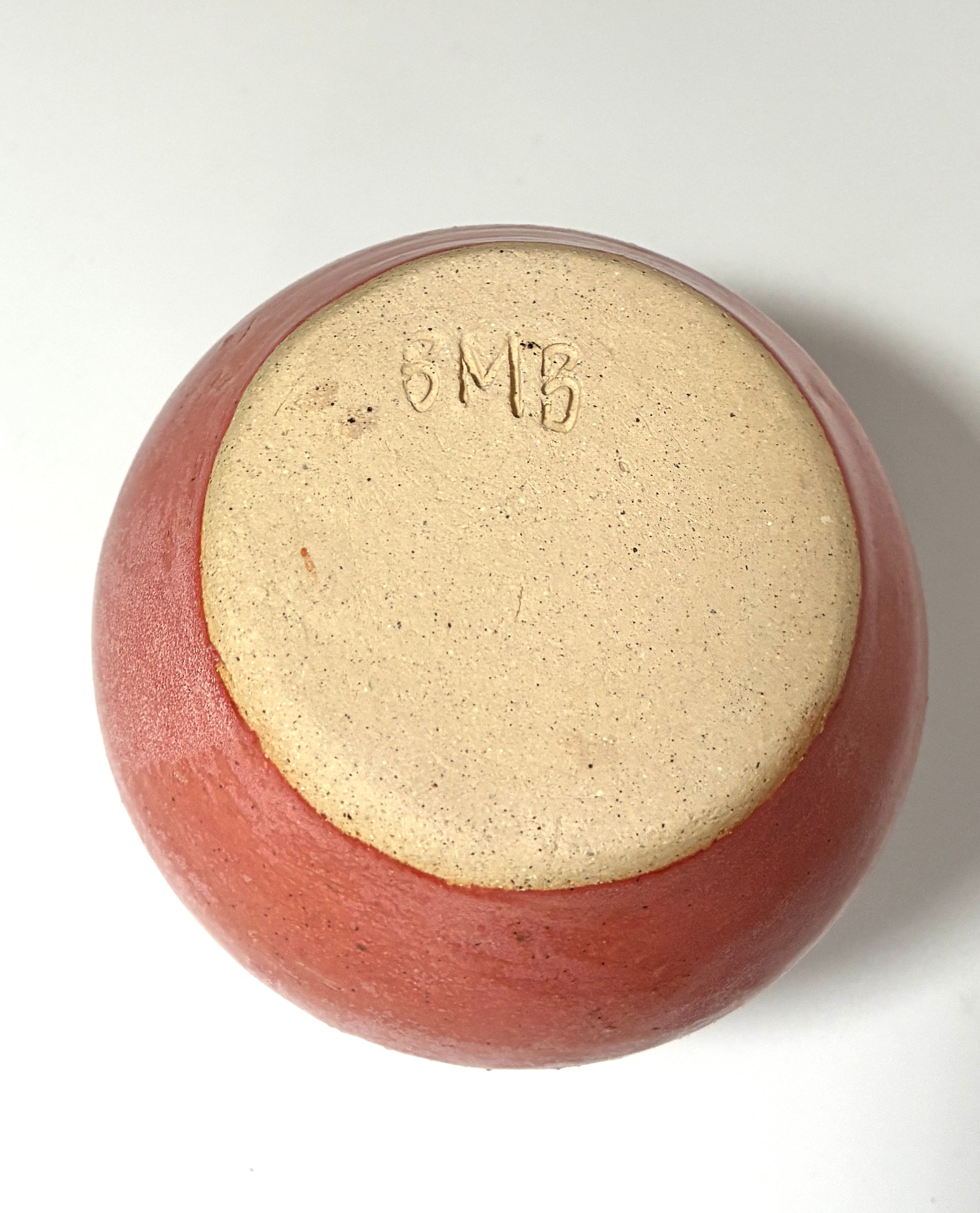 A round, reddish-brown handmade clay vessel from Thyne Goods—called Spam—is shown upside down, revealing an unglazed base with the initials SMB (for Beth Moon Burgess) engraved into the clay.