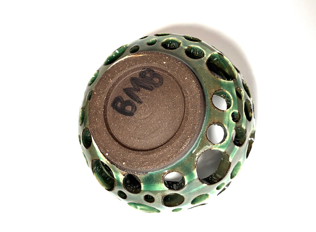 A handmade clay vessel from BMB called “Lettuce,” this round ceramic object is glazed green with many holes and shown upside down on a white background. It features a brown base signed “BMB” in black by the artist.
