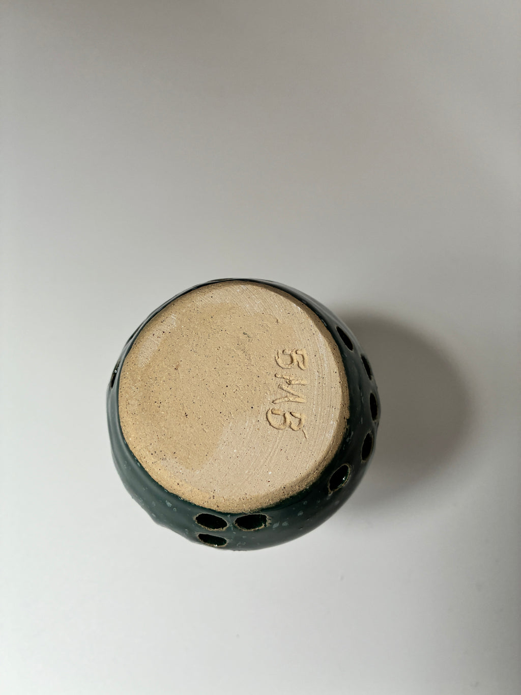 A top-down view of a handmade ceramic pot with a dark green glazed exterior and a natural beige clay base signed with initials BMB