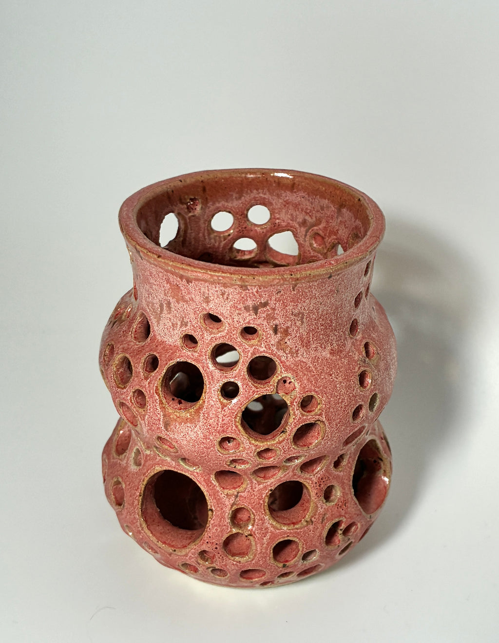 A handmade ceramic vase by BMB, the “Spamalot,” features a glossy pink finish with numerous round holes in various sizes across its surface, reminiscent of a Lonely Worm Farm piece by Beth Moon Burgess, and is photographed against a plain white background.