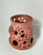 A handmade ceramic vase by BMB, the “Spamalot,” features a glossy pink finish with numerous round holes in various sizes across its surface, reminiscent of a Lonely Worm Farm piece by Beth Moon Burgess, and is photographed against a plain white background.