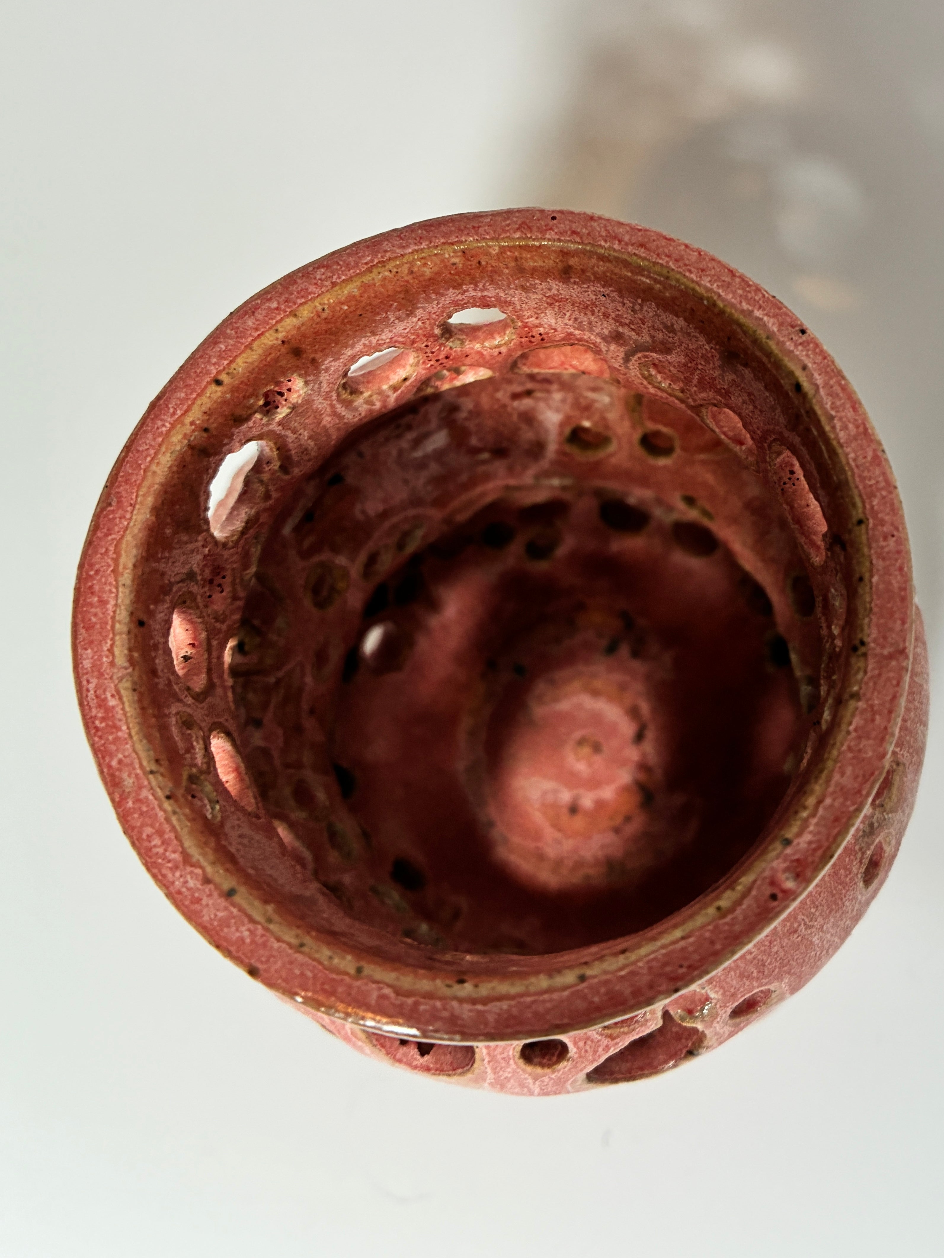 A top-down view of Spamalot—a round, reddish handmade clay vessel by BMB, featuring multiple holes around its rim and sides—displayed on a white surface and evoking the distinctive artistry of the Lonely Worm Farm.
