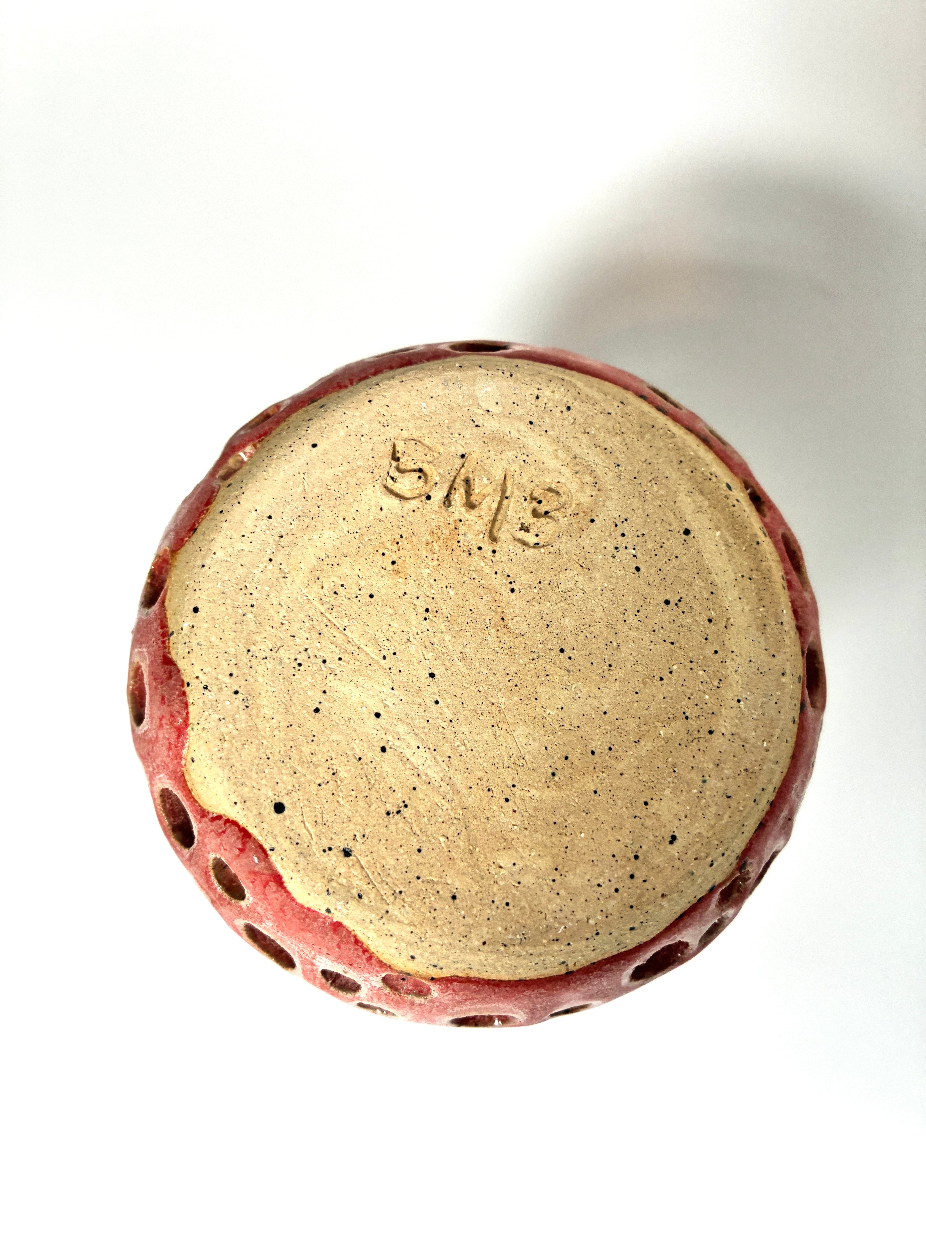 The bottom of Spamalot, a round, beige ceramic vessel with red glaze on the sides and visible holes, shows “BMB” carved into its base. This handmade clay piece by BMB is set against a white background.