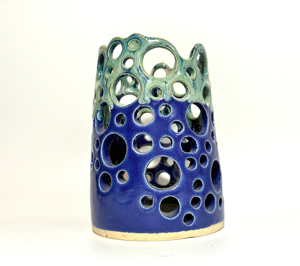 Introducing Widow’s Bay by BMB—a handmade clay vase featuring a blue base and green top, adorned with circular cutouts of varying sizes across its surface. This distinctive piece artfully blends creativity and organic appeal.