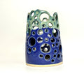 Introducing Widow’s Bay by BMB—a handmade clay vase featuring a blue base and green top, adorned with circular cutouts of varying sizes across its surface. This distinctive piece artfully blends creativity and organic appeal.