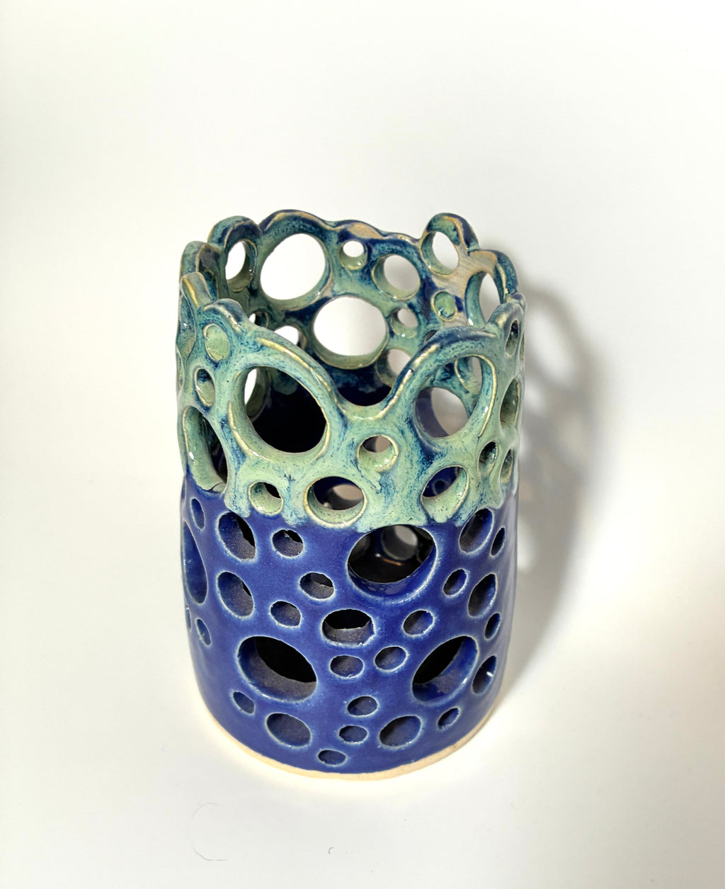 A cylindrical handmade clay vase from the BMB Widow’s Bay collection, featuring circular cutouts and glazed with blue on the bottom half and green on the top, set against a plain white background.