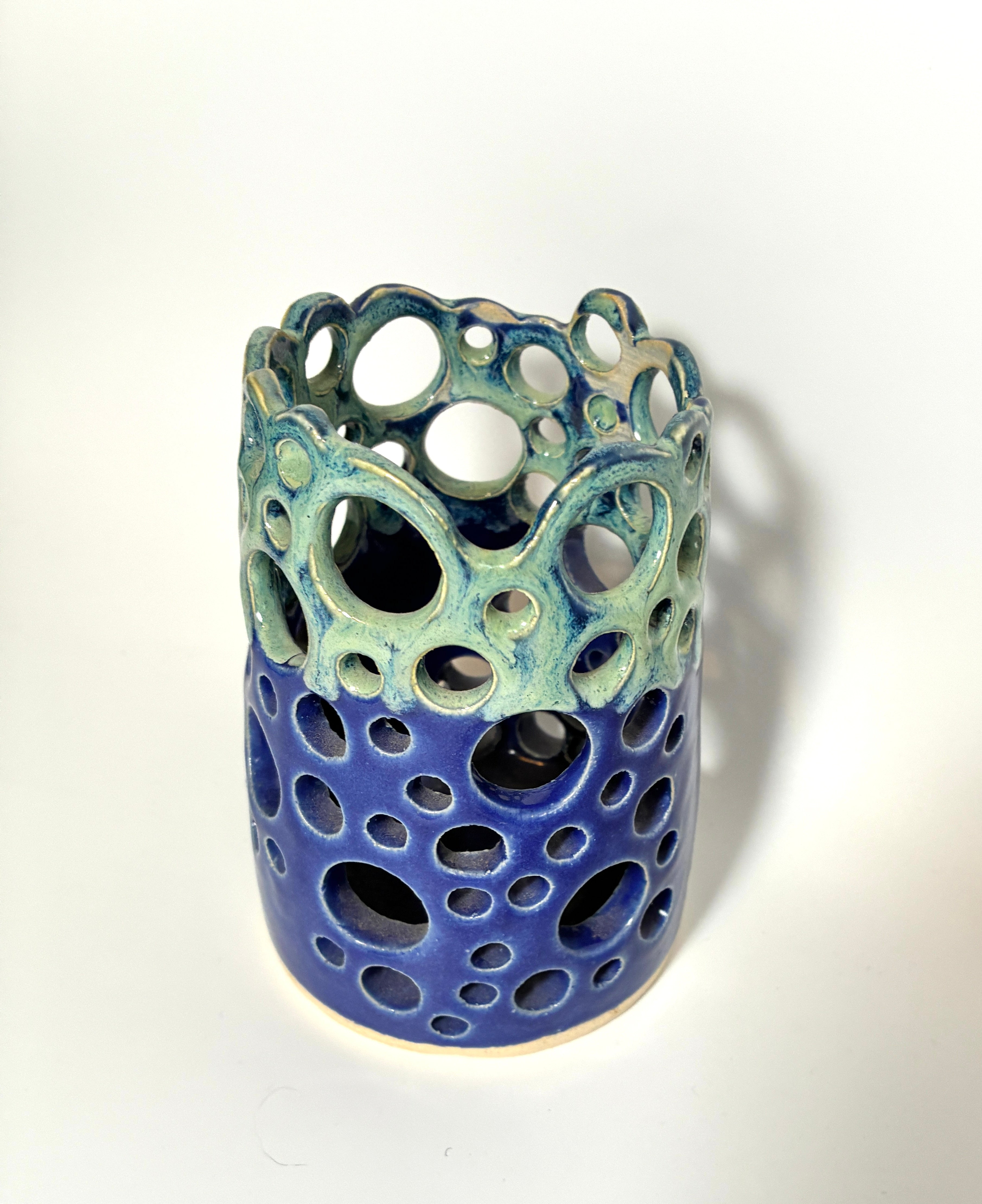A cylindrical handmade clay vase from the BMB Widow’s Bay collection, featuring circular cutouts and glazed with blue on the bottom half and green on the top, set against a plain white background.