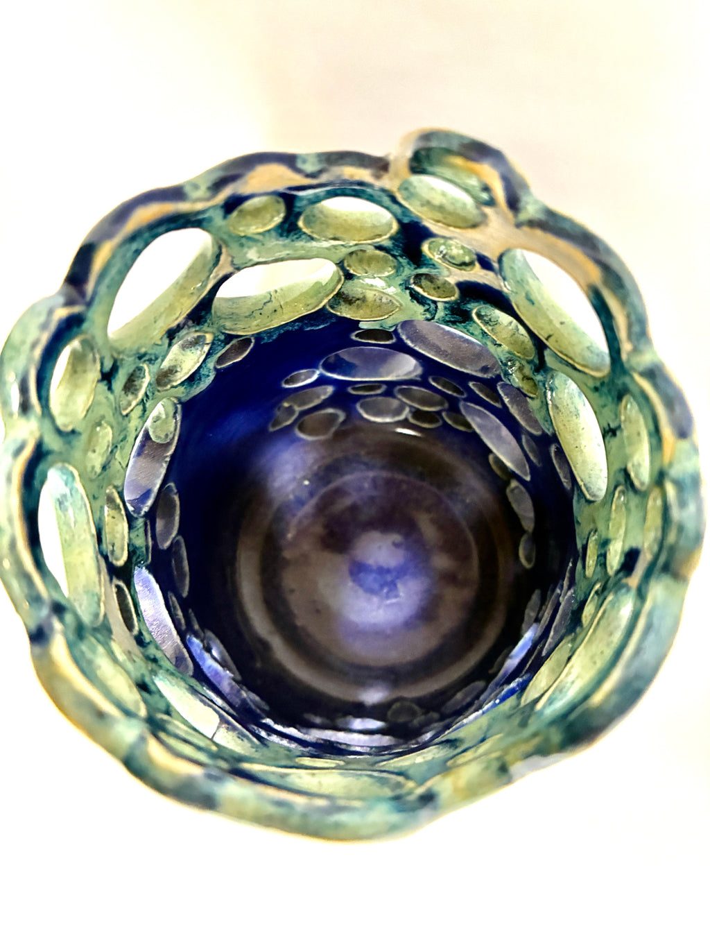 A top-down view of the one-of-a-kind Widow’s Bay decorative glass by BMB, featuring a blue center and a textured, greenish rim with oval cutouts arranged in a circular pattern.