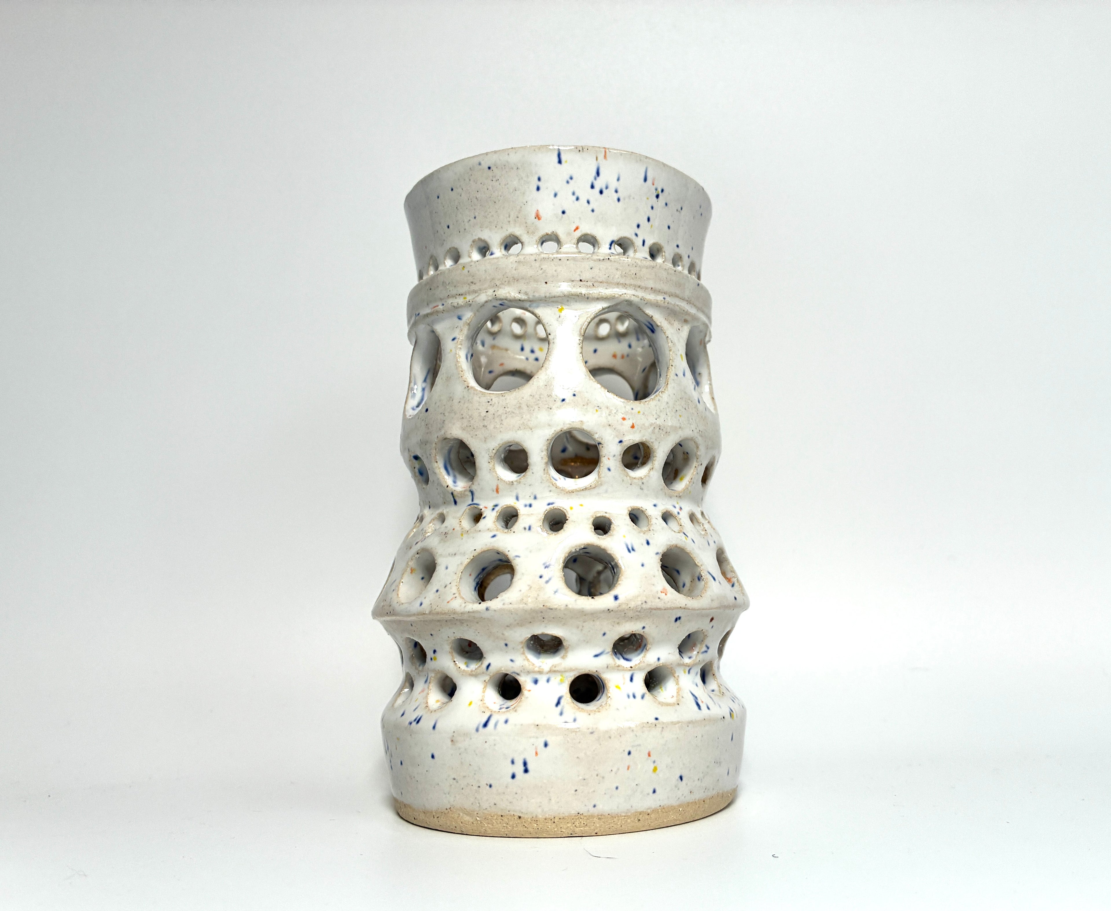 The “Apogee” by BMB is a handmade clay vase in white ceramic, featuring multiple circular cutouts of varying sizes throughout its body. Perfect as a unique home accessory or a stylish vessel for dried flowers, it is displayed on a plain white background.