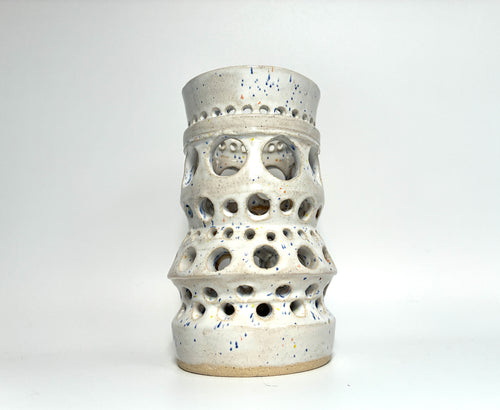 The “Apogee” by BMB is a handmade clay vase in white ceramic, featuring multiple circular cutouts of varying sizes throughout its body. Perfect as a unique home accessory or a stylish vessel for dried flowers, it is displayed on a plain white background.