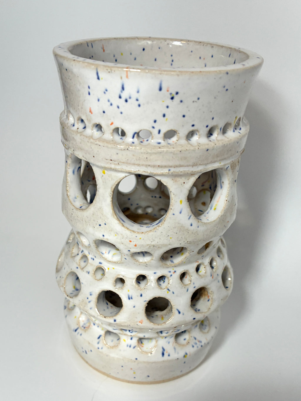 The Apogee vase by BMB is a handmade white ceramic piece adorned with blue speckles and multiple rows of circular cutout holes in varying sizes around its body, making it a striking home accessory.