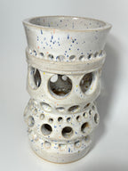 The Apogee vase by BMB is a handmade white ceramic piece adorned with blue speckles and multiple rows of circular cutout holes in varying sizes around its body, making it a striking home accessory.