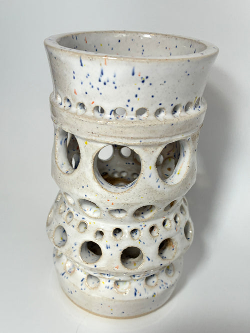 The Apogee vase by BMB is a handmade white ceramic piece adorned with blue speckles and multiple rows of circular cutout holes in varying sizes around its body, making it a striking home accessory.