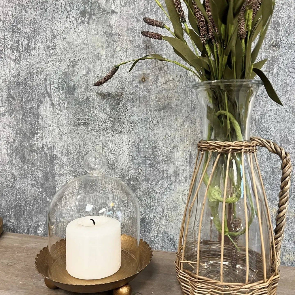 The Cloche Candle Holder with Metal Plate: Gold by Vagabond Vintage features a white candle on a gold tray, paired with a tall vase of dried flowers—ideal for chic home decor against a textured gray wall.