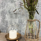The Cloche Candle Holder with Metal Plate: Gold by Vagabond Vintage features a white candle on a gold tray, paired with a tall vase of dried flowers—ideal for chic home decor against a textured gray wall.