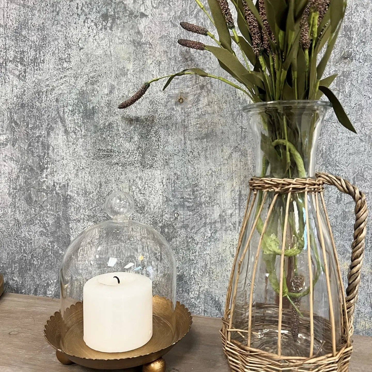 The Cloche Candle Holder with Metal Plate: Gold by Vagabond Vintage features a white candle on a gold tray, paired with a tall vase of dried flowers—ideal for chic home decor against a textured gray wall.