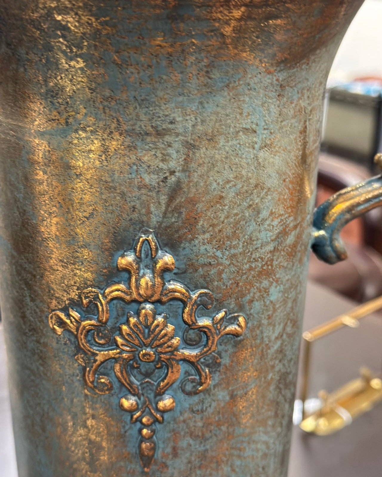 Close-up of the Vagabond Vintage Gold Trophy Vase with Handles, featuring a weathered blue and gold finish and an ornate golden floral pattern in the center. A partial handle is visible on the right side.