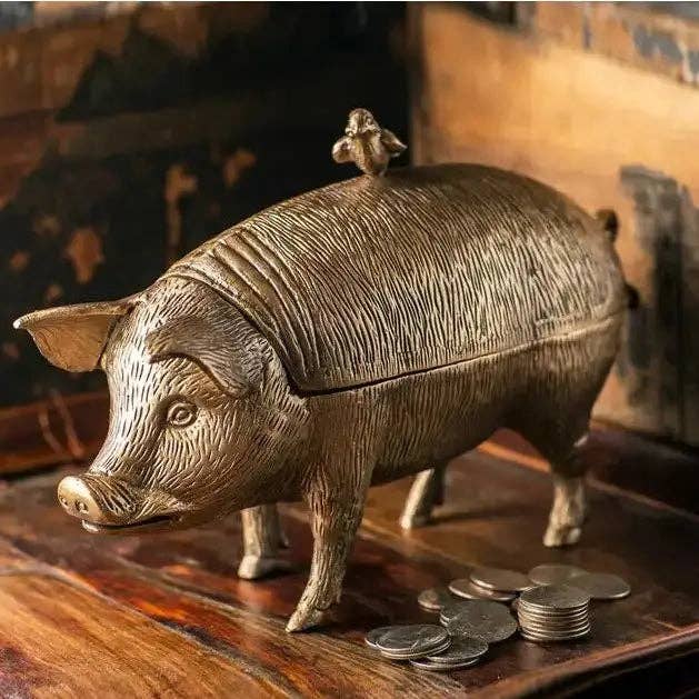The Vagabond Vintage Pig with Bird Storage Box, featuring an antiqued brass finish and coin slot, sits on a wooden surface beside a small pile of coins.